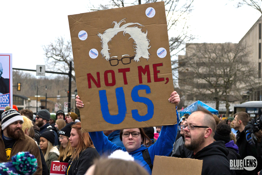 A crowd, including many young people, is gathered to listen to speakers. They are dressed for chilly weather, and many wear knit caps. One person in a blue fleece jacket and earrings holds up a hand-painted sign with an image of Bernie Sanders’ trademark unruly white hair over colored blue and red lettering that reads, “Not me, us.”