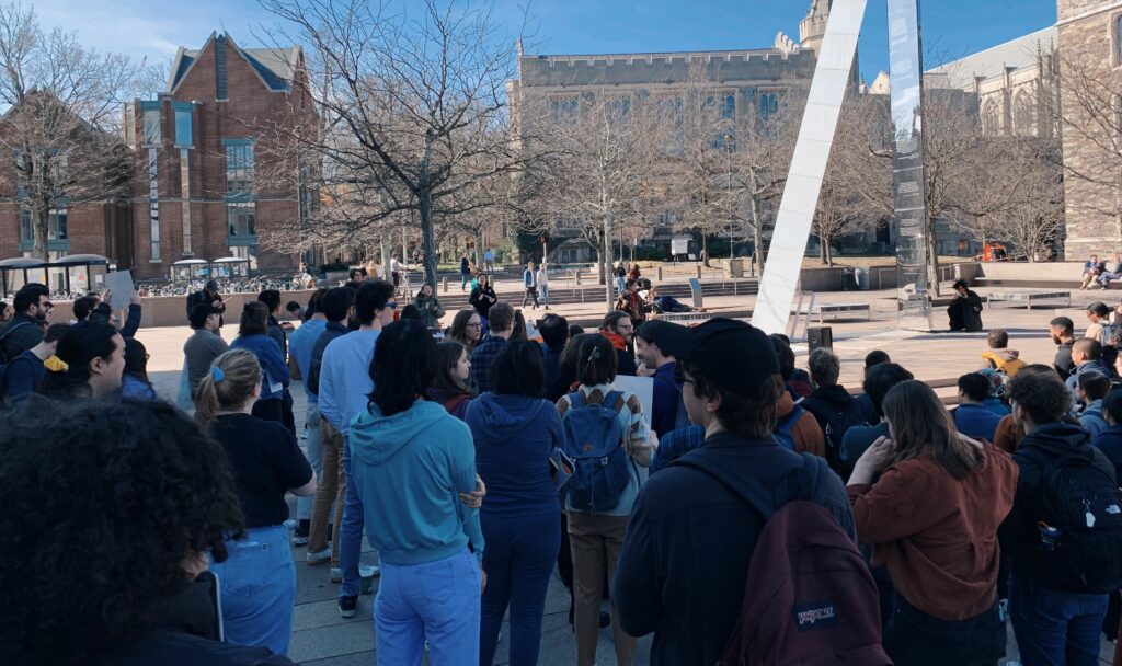 A photo of a large group of people gathered in a courtyard of Princeton University’s campus. Some are holding signs but the viewer cannot read what’s written on them because people are facing the opposite direction of the camera. Trees are barren, marking the winter months.