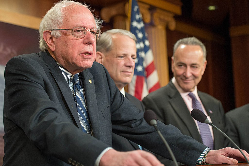 Bernie Sanders speaks at a podium in front of an American flag. He is flanked by Senate Democrats, including Chuck Schumer, who looks on smiling.