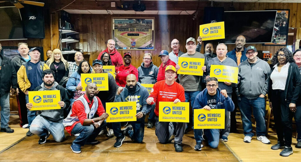 About 20 workers stand in a paneled room holding signs reading "Members United."