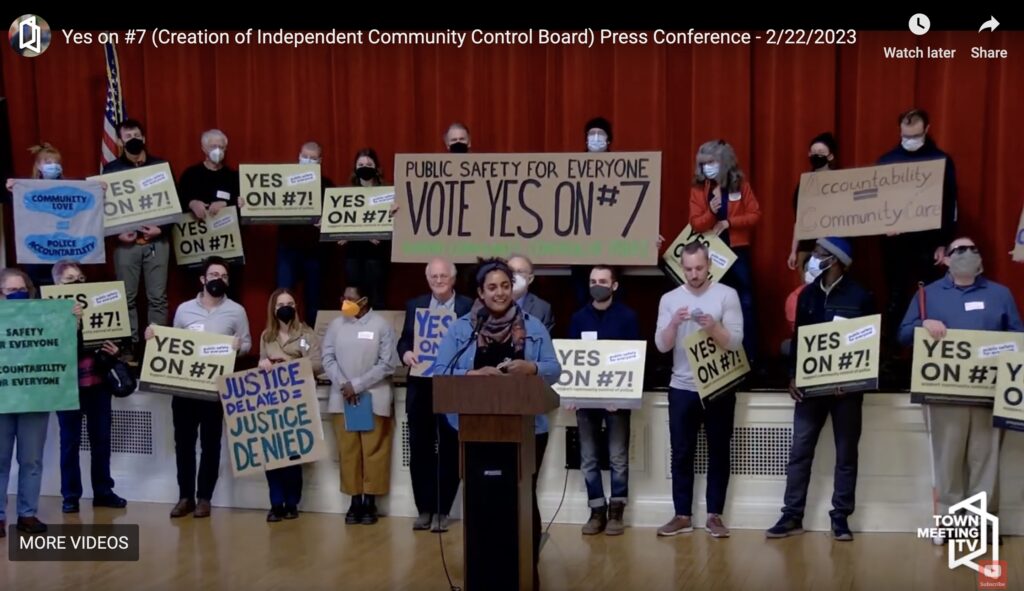 A screen shot of a video shows about twenty-one people standing in an auditorium, shoulder to shoulder, facing the camera, and holding various signs. One person, Jess Laporte, stands in the front at a podium, smiling and speaking into a microphone. Majority of the signs read “YES ON #7!” A large sign held up by two people says “PUBLIC SAFETY FOR EVERYONE - VOTE YES ON #7.” Additional signs say “Accountability = Community Care,” another “SAFETY FOR EVERYONE - ACCOUNTABILITY FOR EVERYONE,” “COMMUNITY LOVE = POLICE ACCOUNTABILITY,” and “JUSTICE DELAYED = JUSTICE DENIED.” Majority of the people are wearing either surgical masks or N95’s. In the video’s corner, a logo says “TOWN MEETING TV” and at the top, a video title says “YES ON #7 (Creation of Independent Community Control Board) Press Conference - 2/22/2023.”