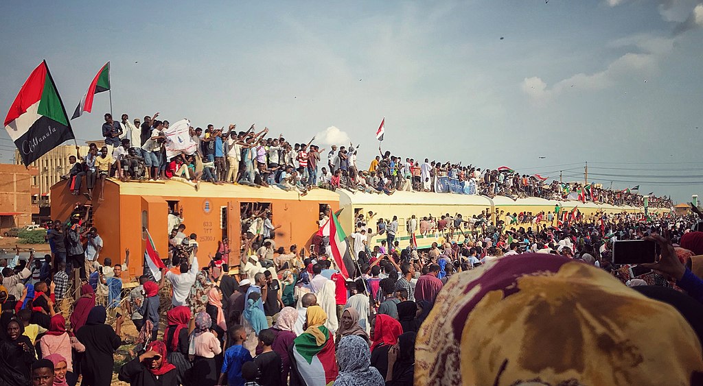 Many Sudanese people stand on top of, in, and around about six cars of a yellow train. Some wave the Sudanese flag. In the forefront are several people watching and taking photos of the train.