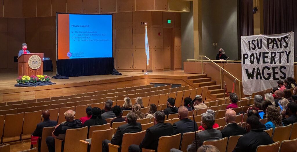 The ISU President Terri Goss Kinzy speaks at a podium to an audience in an auditorium. A large monitor stands to the President’s right, currently displaying a slide titled ‘Private support.’ On the side is a large white banner draped and in black text says, ‘ISU PAYS POVERTY WAGES.’