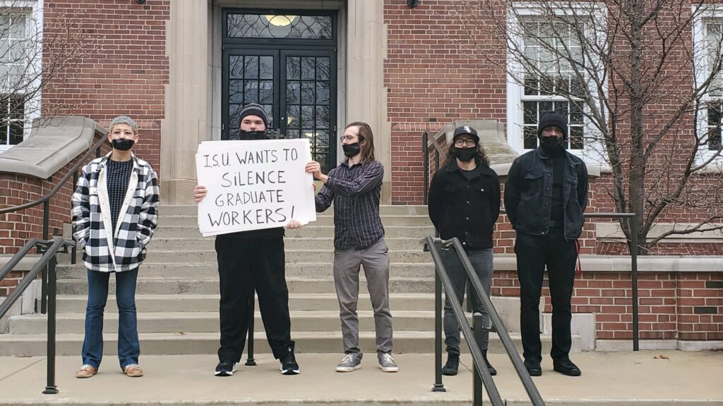 Five people stand uniformly in a row in front of a university brick building with their mouths taped shut by black tape, facing straight ahead. Two people hold a sign that says ‘ISU WANTS TO SILENCE GRADUATE WORKERS!’ Two people stand with hands behind their backs, and the last person has their hands in their sweater’s pockets.