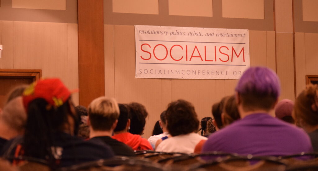 Multiple people are seated and facing ahead, with the camera capturing the backs of their heads. The wall they are facing has a large white banner that reads ‘revolutionary politics, debate, and entertainment. SOCIALISM. SOCIALISMCONFERENCE.ORG.’ The word SOCIALISM is in red. It seems like attendees are watching speakers.