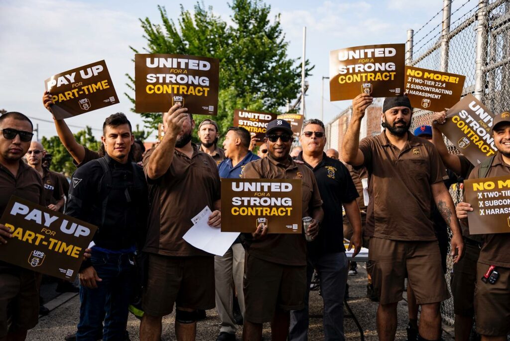 15 people in brown uniforms, Teamsters workers at UPS, stand outside a fence holding signs that say "United for a Strong Contract," "Pay Up for Part Time."