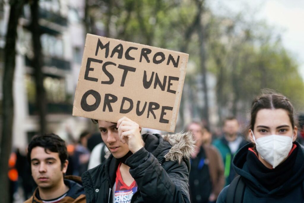 Three young demonstrators at the the 11th inter-union day of action against pension reform on April 6, 2023 in Paris, France. The center demonstrator is holding  a sign stating "Macron is a scumbag".