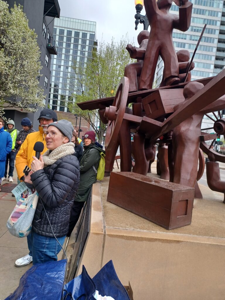 A person in winter dress holds a microphone in front of Chicago's Haymarket memorial, a brown statue of abstractly rendered people building a wagon together.