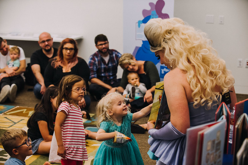 A drag queen with big blond hair in a purple dress with matching fingerless opera gloves that go from the wrists to the elbow is reading a large colorful book to the kids. Some are sitting on a rug in the center of the room and two children are standing close to the drag queen. One is wearing a red and white horizontal striped shirt and is looking curiously at the book while the other child is in a green dress, smiling and pointing with their left hand at something in the book while their right hand is stuck inside a bag of chips. A number of adults are sitting off to the side and some of them are holding smaller children sitting in their laps.