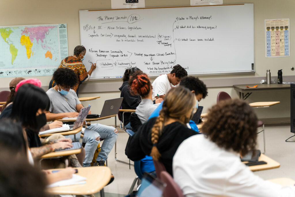 A group of students sits at desks with laptops and papers reading a white board on which a teacher writes a lesson about the Plessy v. Ferguson Supreme Court Decision.