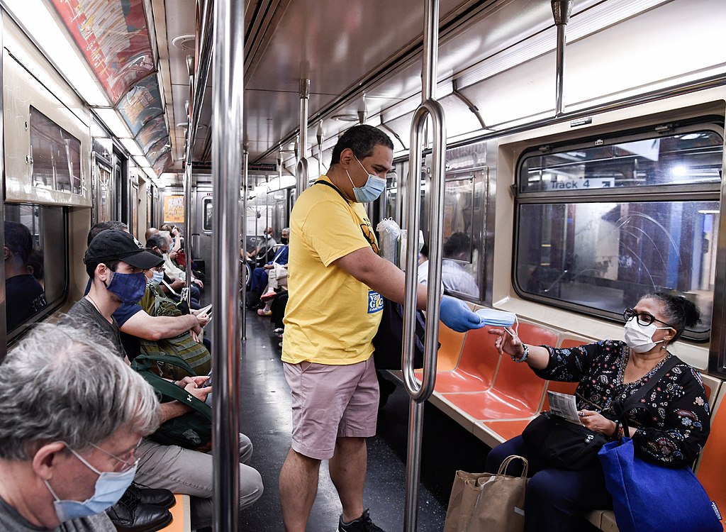 A volunteer with the MTA Mask Force wearing a bright yellow shirt, a blue mask, and matching blue hospital gloves is handing a free mask to a person sitting down in a subway car. Several other people are sitting on the train wearing masks.