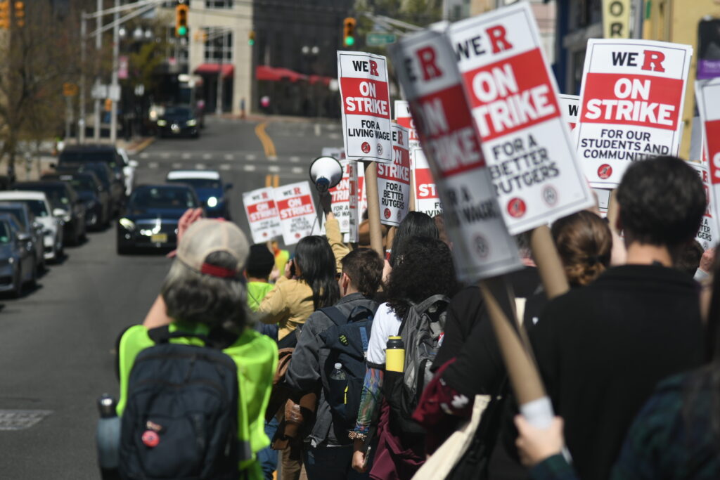 A tight group of dozens of marchers proceed down a city street carrying signs reading "We R on strike for a better Rutgers." The shot is taken from behind the march.