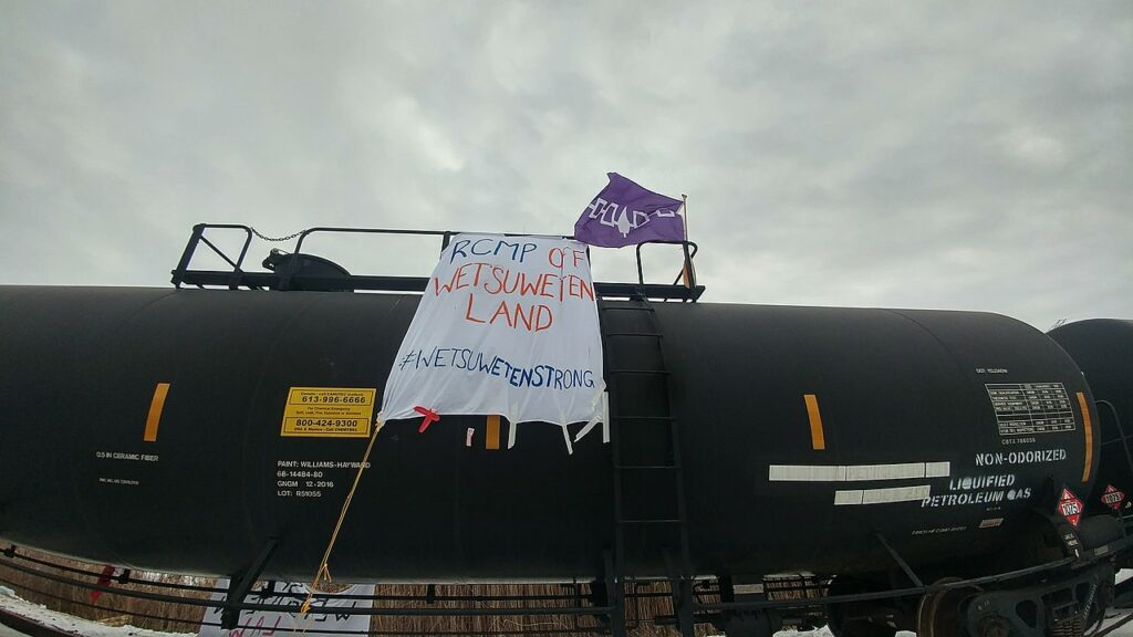 A white banner painted with the words "RCMP OFF WET’SUWET’EN LAND" and the hashtag #WETSUWETENSTRONG in blue and red lettering drapes a black tank, with a Haudenosaunee flag, that is purple with white design on it, flying on railings on top of the tank. The tank has various lettering on it describing its contents, such as ‘NON-ODORIZED LIQUIFIED PETROLEUM GAS’ and product numbers.