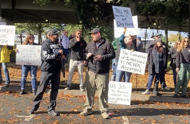 A dozen right-wing protesters wearing winter coats stande on a parking lot strewn with fall leaves. They hold signs reading "Save God's Children," "Tell Me that this is not Child Abuse," "The Corruption of Innocence is NEVER Acceptable," and "OUR Job to protect children from EXPLOITATION and HARM.
