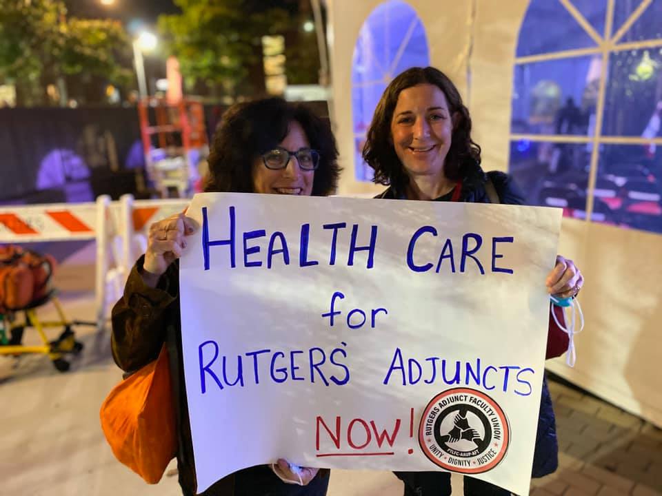Two women standing in front of a building at night hold a white sign with blue lettering reading Healthcare for Rutgers Adjuncts Now!