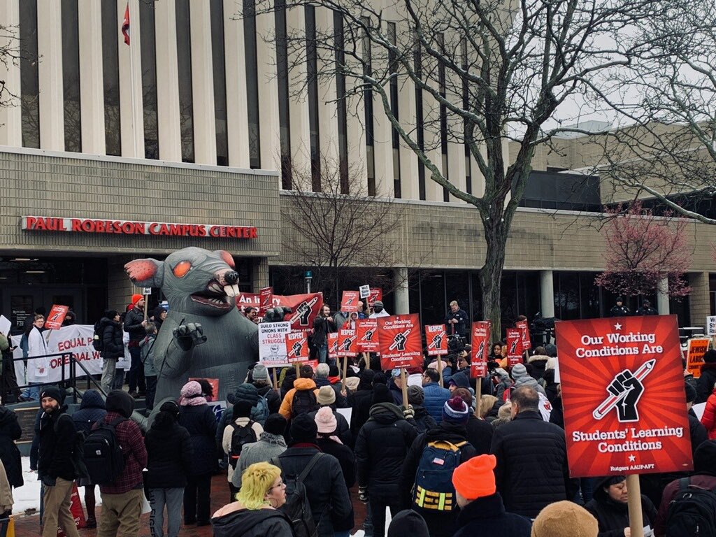 A crowd of demonstrators are gathered before the Robeson cultural center at Rutgers university holding red signs reading Our working conditions are students' learning conditions. At the front of the assembly is a person in a giant rat costume.