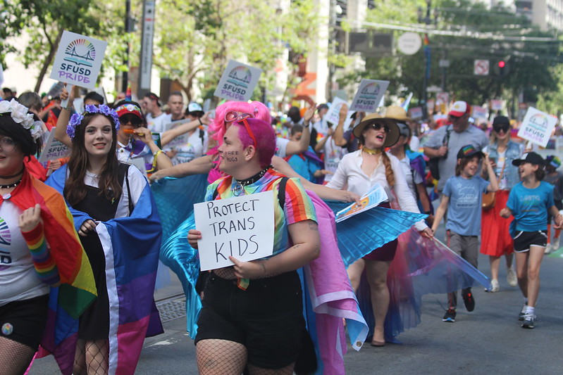 Young people with colorful hair and wearing pride flags, including a pink, white, and blue trans pride flag, and flowers in their hair, march with hundreds of others in SFUSD pride. One marcher holds a small sign reading "PROTECT TRANS KIDS."