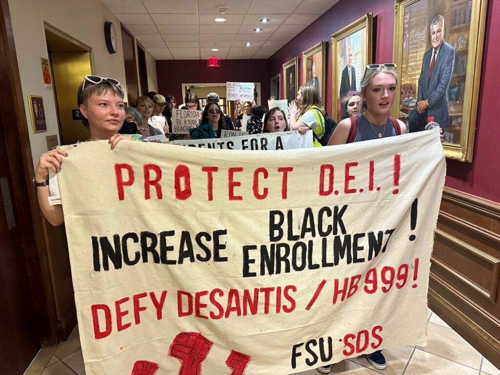 Rows of students march in a hallway at Florida State University administration building flanked by portraits of white, male university presidents. They carry banners and signs reading "PROTECT D.E.I! INCREASE BLACK ENROLLMENT! DEFY DESANTIS/HB999!" A banner has images of red raised fist and Identity the marchers as FSU-SDS.