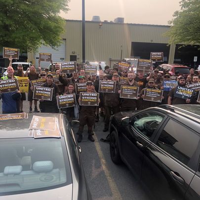 A group of about 40 UPS workers wearing brown are gathered in a parking lot holding signs reading "United for a Strong Contract."