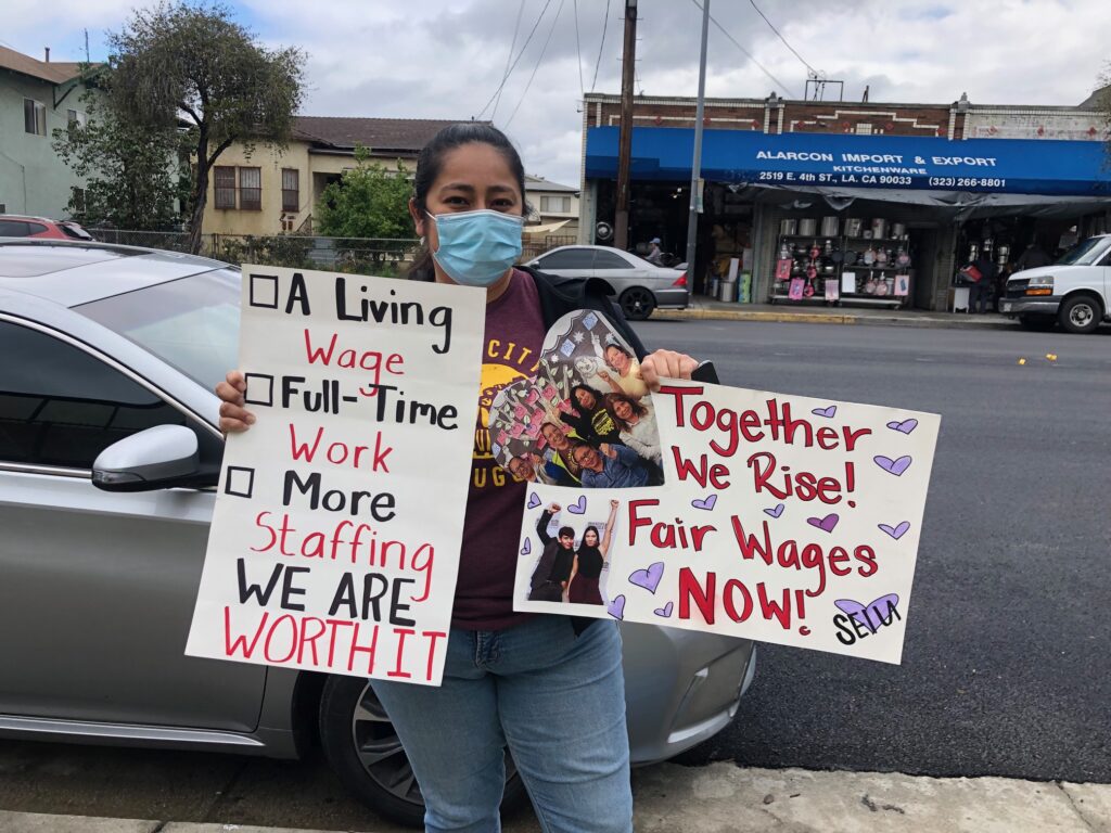AN SEIU Local 99 striker holding up two posters while demonstrating. The first has three check boxes next to three demands, respectively, a living wage, full time work, more staffing with the slogan, “We are worth it.” The second poster reads: “Together we rise” Fair wages now.”