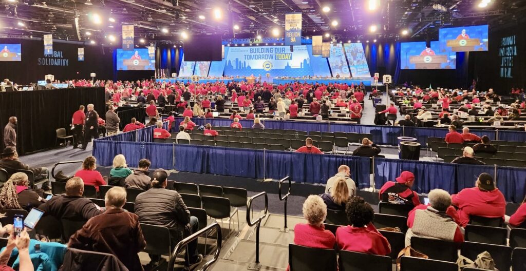 A photo of a large meeting in a large convention room. Many rows of seated individuals face a stage that has speakers and a panel. The platform is backed by large lighted screens showing various photos of UAW strikes. The main screen reads ‘BUILDING OUR TOMORROW TODAY’ and shows the UAW logo. Other screens show live filming of the person speaking at a platform on the stage. Various flags adorn the room’s ceiling, indicating the region of that part of the room. The flags legible in the photo read, REGION 9, REGION 9A, and REGION 4. Most of the people in the photo are wearing red shirts.