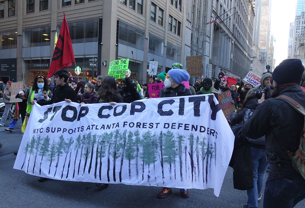A large gathering of environmental and anti-racism anti-police protestors marching through NYC by several buildings including a Starbucks in the left corner of the photo are holding signs. A prominent sign with a white backdrop upfront reads “STOP COP CITY” in large black writing, followed by the line “NYC Supports Atlanta Forest Defenders” in smaller black writing, with a row of green trees taking up about half the sign. Other signs in the background of the photo include a green sign reading “Defend the forest everywhere stop cop city,” a cardboard sign reads “Defund the police, invest in black communities,’ another cardboard sign reads “Police do not protect people, they protect property,” and another sign reads “Uproot the system.”