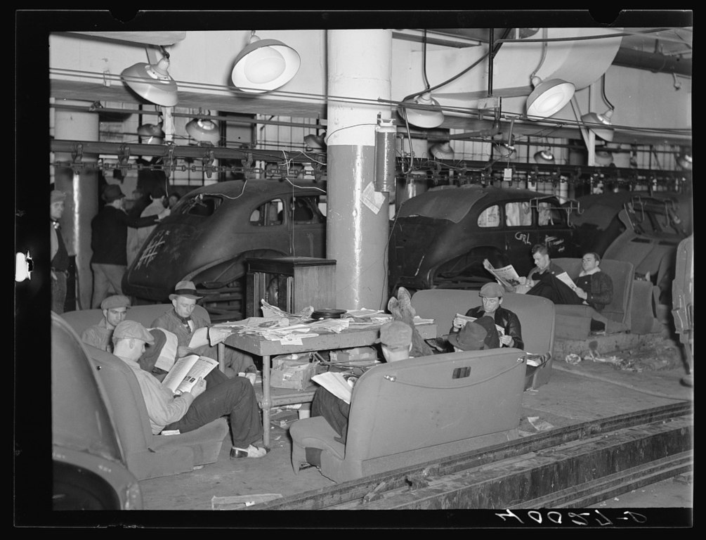 A black and white photo of an autofactory. In the forefront, eight men are sitting in various automobile seats reading newspapers. The seats are placed on the ground and have yet to be installed in the cars. In the background, an assembly line shows 3 vehicles stalled in the process of being made. There is a group of people standing and talking behind the cars. The ceiling is lined with factory lighting.