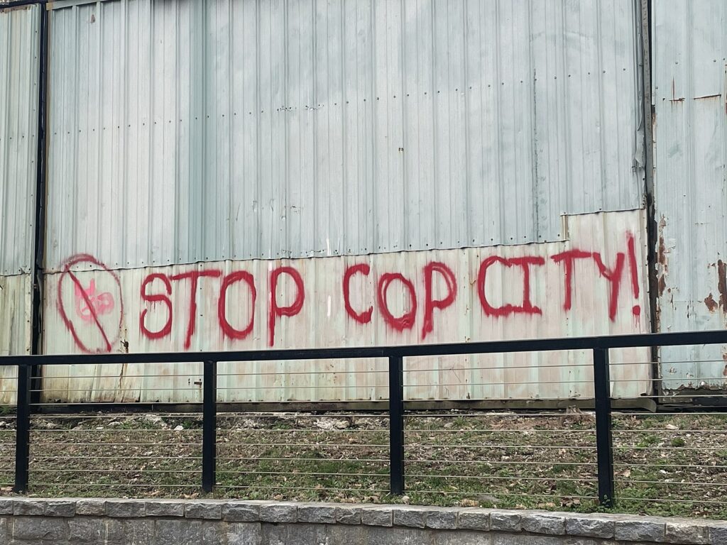 The message "Stop Cop City" is painted in red graffiti on a white building located behind a fence along the Proctor Creek Greenway Trail in Atlanta, GA.