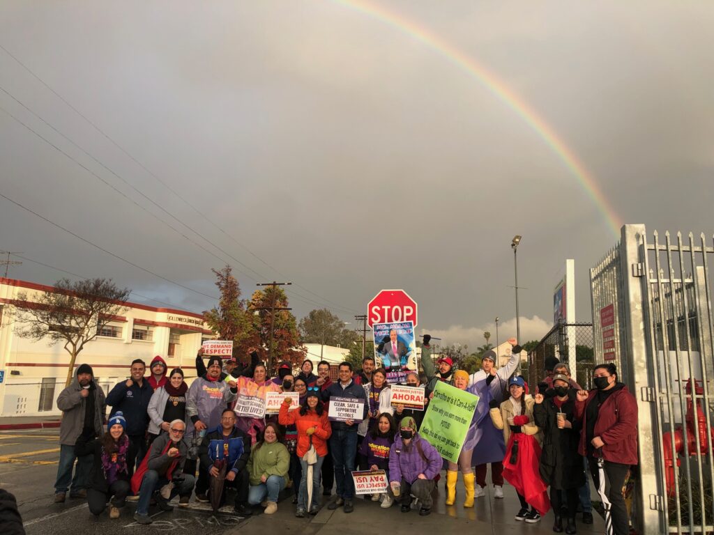 A group of about twenty UTLA and SEIU members and supporters gathered holding posters, ad pickets, beneath a rainbow following rainfall.