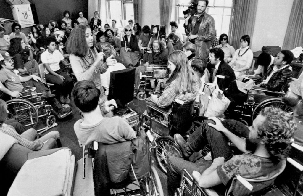 About 50 people, many in wheelchairs, gather around a young person speaking. Media cameras are trained on the speaker in this black-and-white image of the historic 1977 HEW 504 sit-in.
