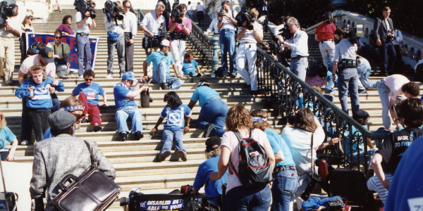 About 20 demonstrators, many in blue shirts, climb the steps of the U.S. Capitol while media record the scene. Other protesters, some in wheelchairs, stand by.