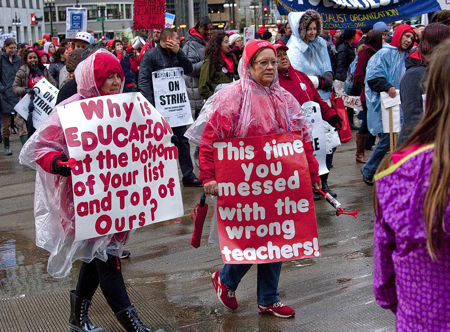 Teachers in rain gear demonstrating on the street with banners and picket signs. One figure in the foreground carries a sign saying, “Why is education at the bottom of your list and top of ours?” The sign of the second figure in the foreground reads, “This time you messed with the wrong teachers!”
