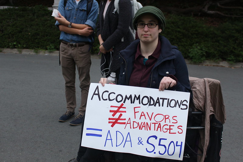 A person in a green hat, black glasses, and blue coat sits in a wheechair holding a sign reading "Accomodations ≠ Favors ≠ Advantages =ADA & 504." Two people stand behind.