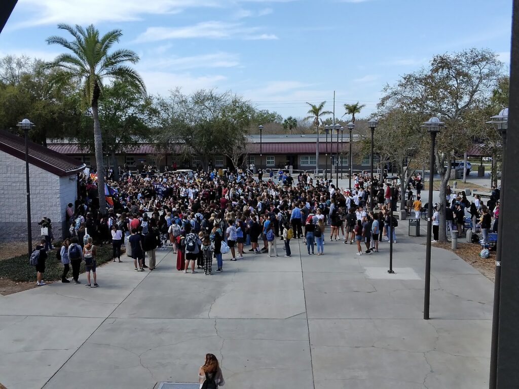 A large group of high school students casually dressed from Palm Harbor University High School in Palm Harbor, Florida are gathered close together on the pavement after walking out of the school in protest of the state's "Don't Say Gay" bill. Some palm trees are lined around the pavement where the students are gathered.