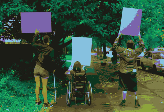 Three people are shown from behind holding up signs toward oncoming traffic along a wooded street. One holds a cane, one is in a wheelchair, and the third has no assistive devices.