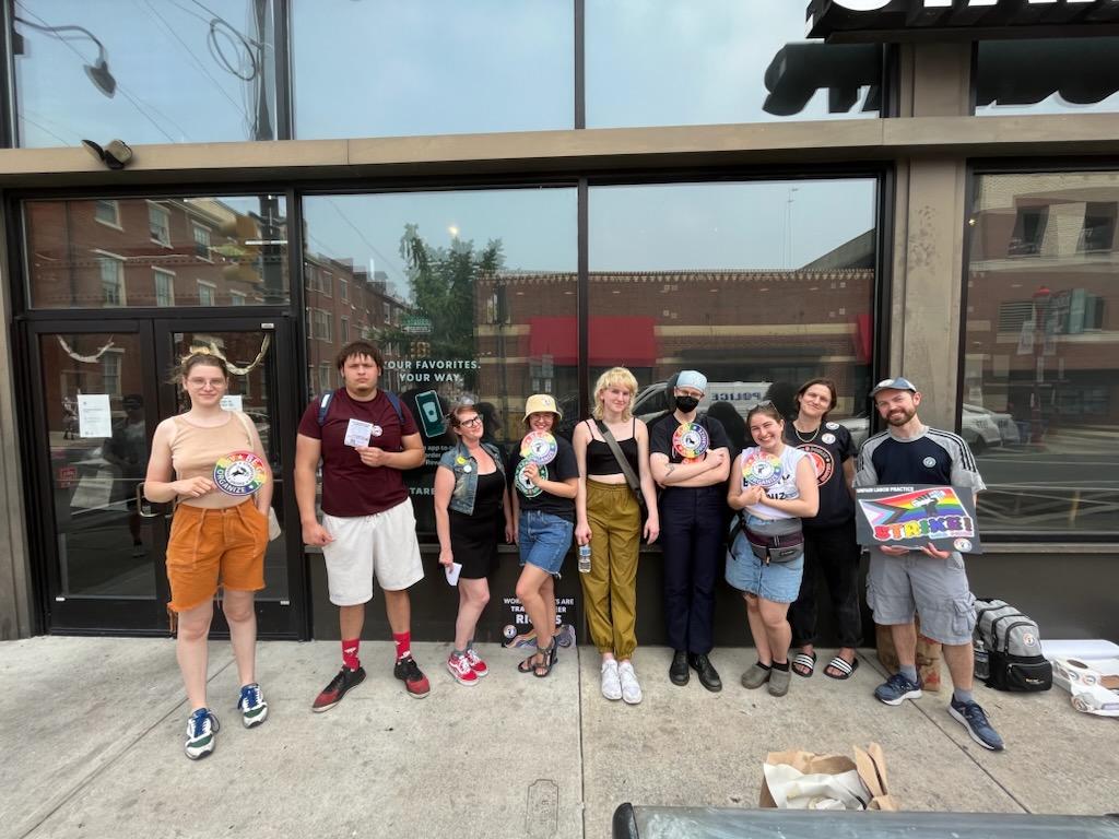9 people stand in front of a storefront holding rainbow signs