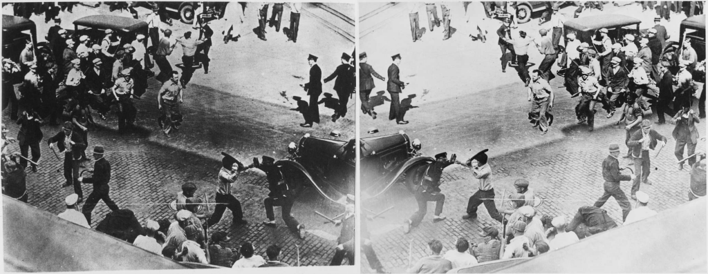 Black and white photograph taken from above shows a man holding a metal pipe mid-swing as a uniformed police officer shields his face with an outstretched hand; in the foreground, just visible over the roof of a truck is the back of an officer who has tackled someone to the ground; in the background, a crowd of pipe-wielding men advances down a cobblestone street and another man moves toward a cop who is hoisting a nightstick overhead.