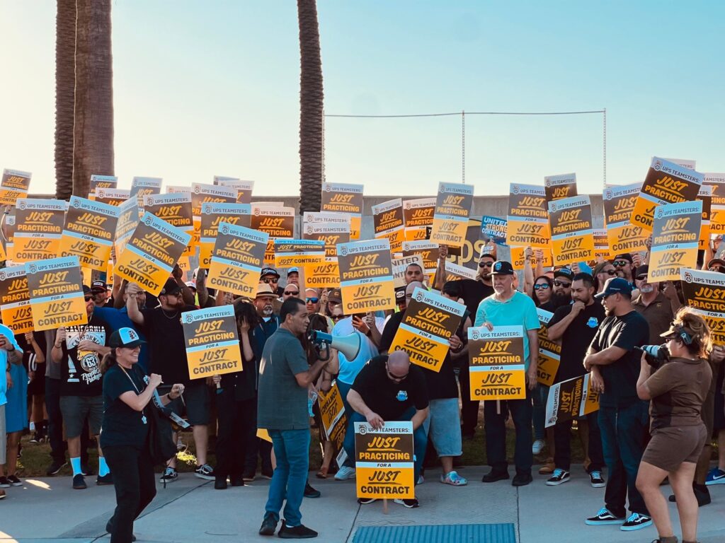 A racially and gender-diverse crowd of workers, some wearing the brown UPS uniform, hold up yellow and brown picket signs (the colors of the UPS company logo); the picket signs read “UPS TEAMSTERS JUST PRACTICING FOR A JUST CONTRACT.”