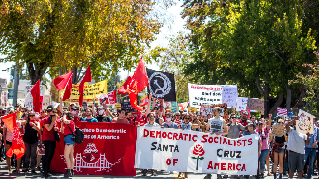 DSA members stand at the front of a crowd of protesters waving red flags and holding two banners that read “San Francisco Democratic Socialists of America” and “DEMOCRATIC SOCIALISTS SANTA CRUZ OF AMERICA.”