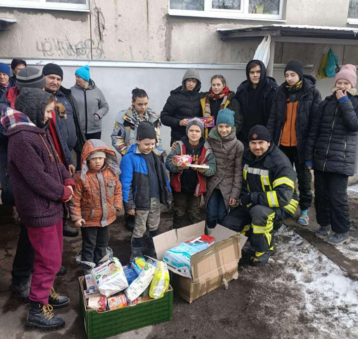 About 15 men, women and children pose with two cardboard boxes of packaged aid. The people are bundled up against the cold, and one boy holds up a couple of small items from the aid package.