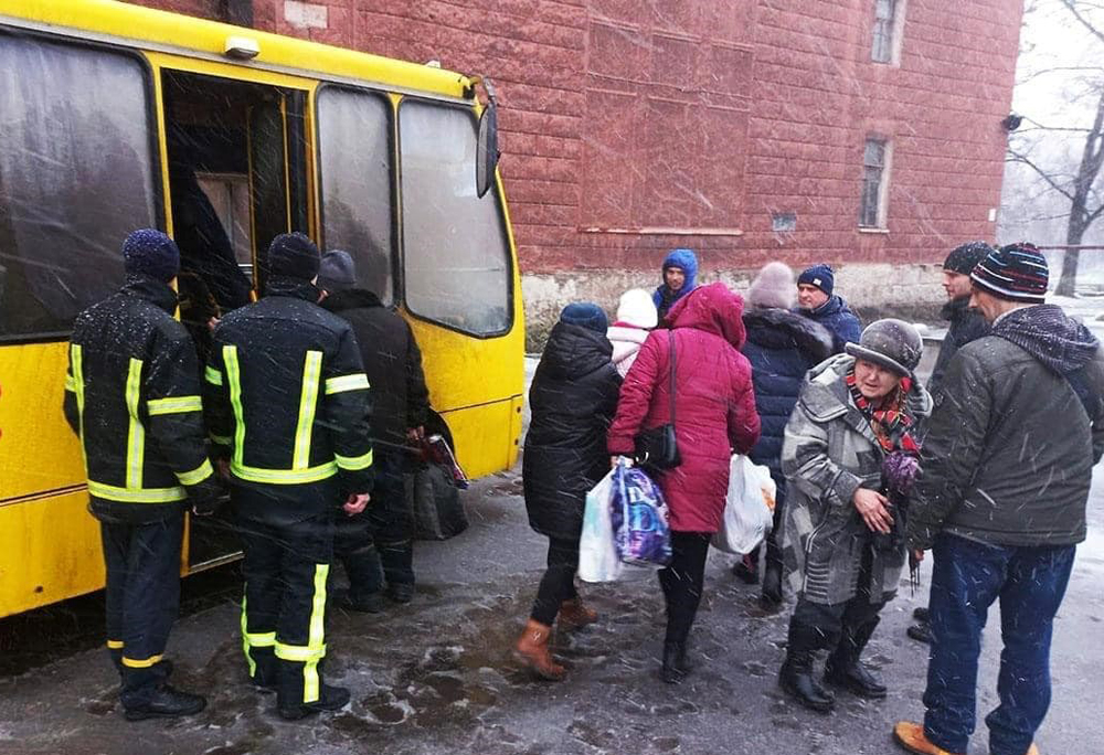 About ten adults holding luggage and bags mill around outside a yellow bus. Two evacuation workers in reflective clothing watch people board the bus. All are bundled against the cold, and there is snow on their shoulders, hoods, knit caps.