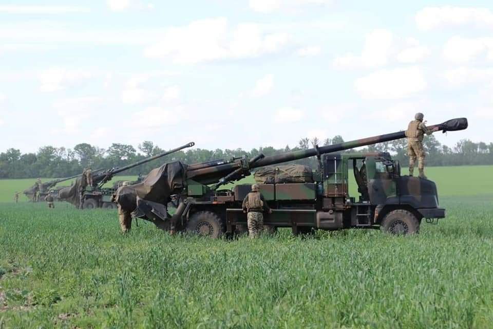 A row of three howitzers, mounted on heavy trucks, face the same direction in a green field. These cannons, which measure ten to twelve meters in length, tower over the soldiers who are tending them.
