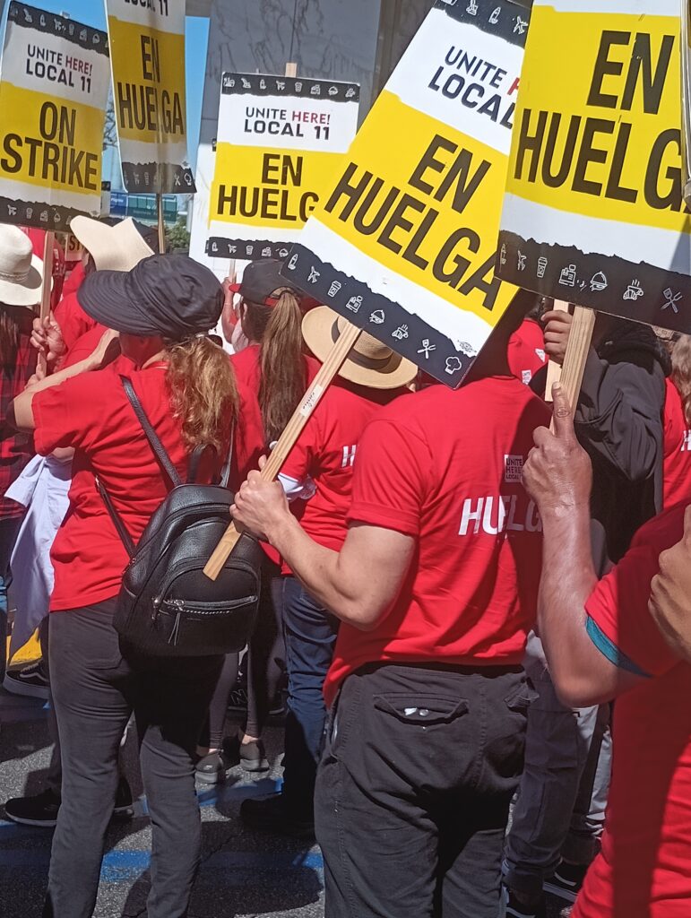 A crowd of people in red shirts carry yellow and white signs reading "en huelga" (on strike).