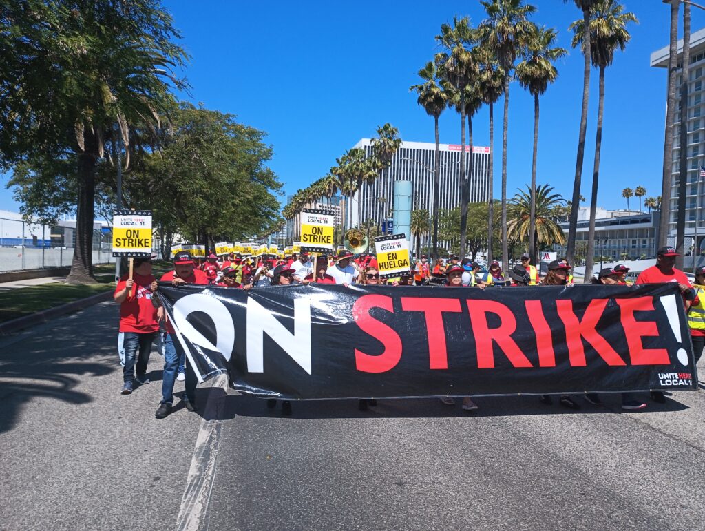 Workers march behind a black red and white banner reading "on strike!" along a palm-tree lined city street in Los Angeles.