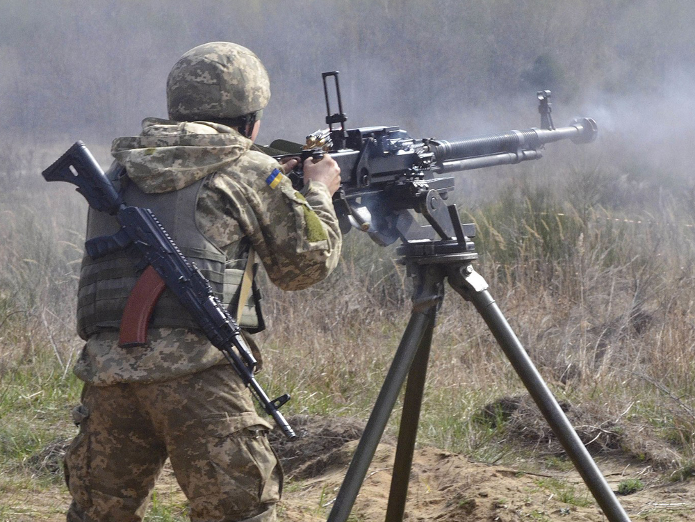 A soldier in camouflage stands and fires a large machine gun across a field. The gun is  mounted on a tall tripod, and the soldier, their back turned to the camera, has another automatic rifle slung over their back.