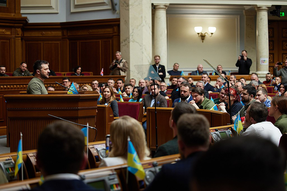 President Zelensky in an olive-drab T shirt addresses the seated members of parliament in a medium long shot. Zelensky is in profile, looking right, and the room features off-white marble columns and a lot of dark tooled wood.