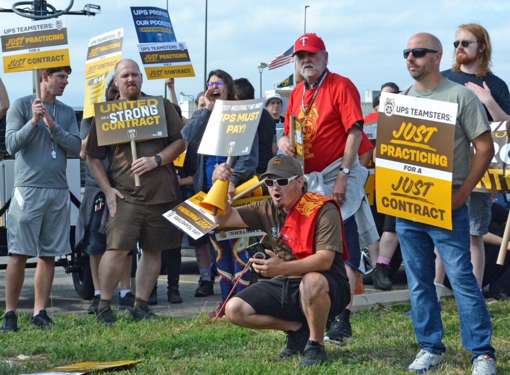 About a dozen of women and men are clustered together; one holds a megaphone and one is shouting. They carry signs reading just practicing for a just contract, united for a strong contract, ups must pay.