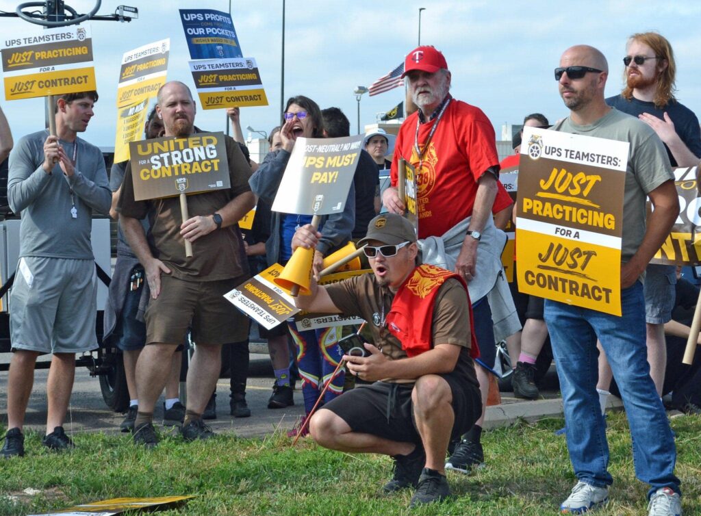 About a dozen of women and men are clustered together; one holds a megaphone and one is shouting. They carry signs reading just practicing for a just contract, united for a strong contract, ups must pay.