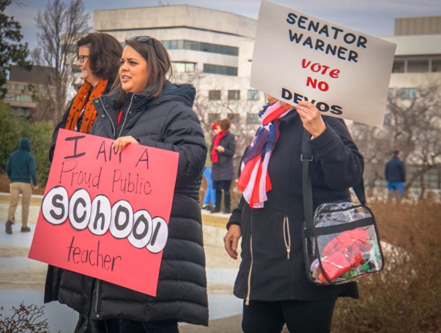 Three people in winter coats stand in front of tall white buildings holding signs reading I am a proud public school teacher and Senator Warner vote no Devos