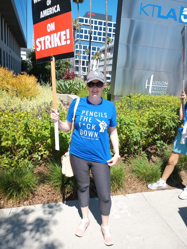 A woman in blue holds a picket sign reading strike! Her shirt reads: Pencils the f*** down.